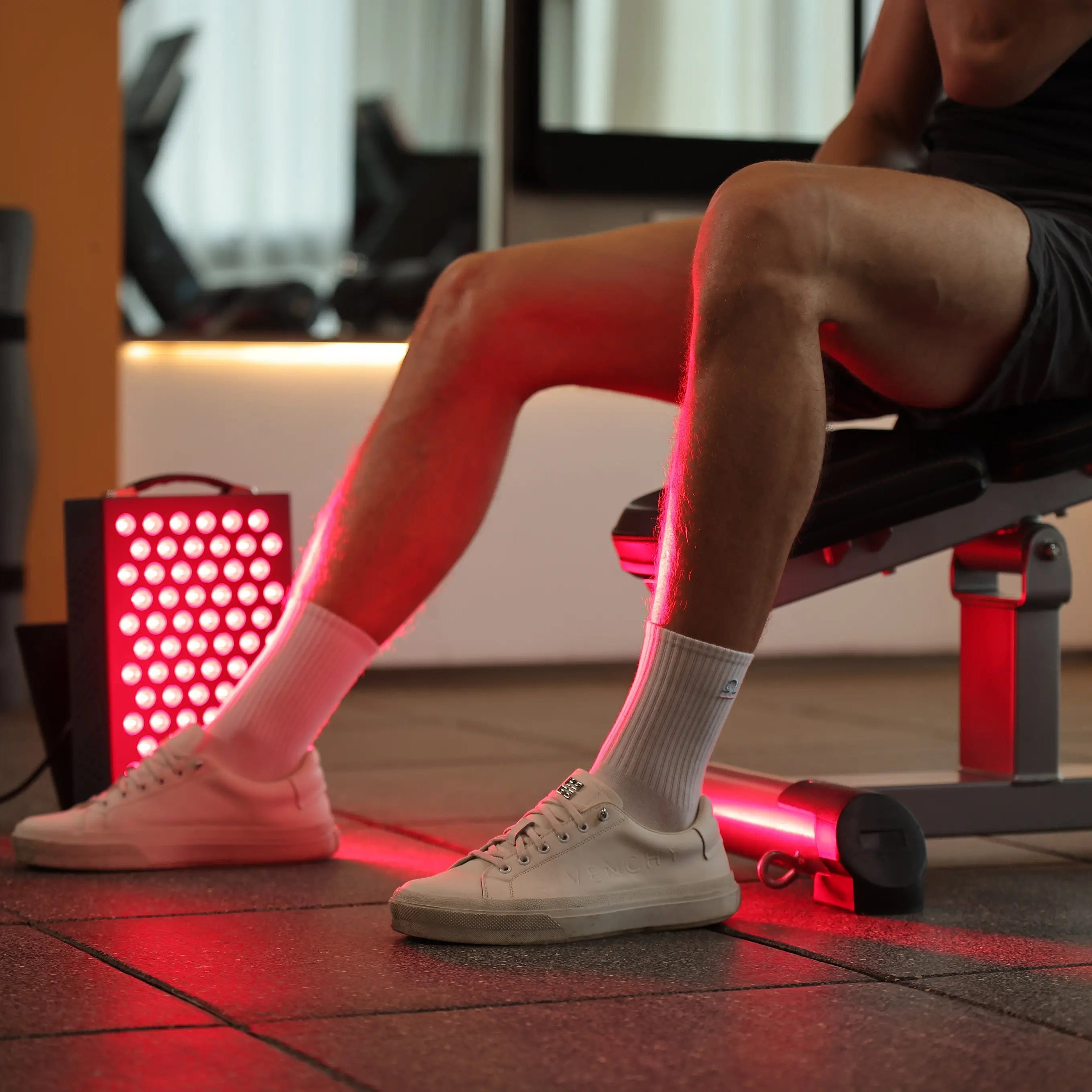 Athlete using YouLumi red light therapy panel for lower-leg recovery beside a gym bench
