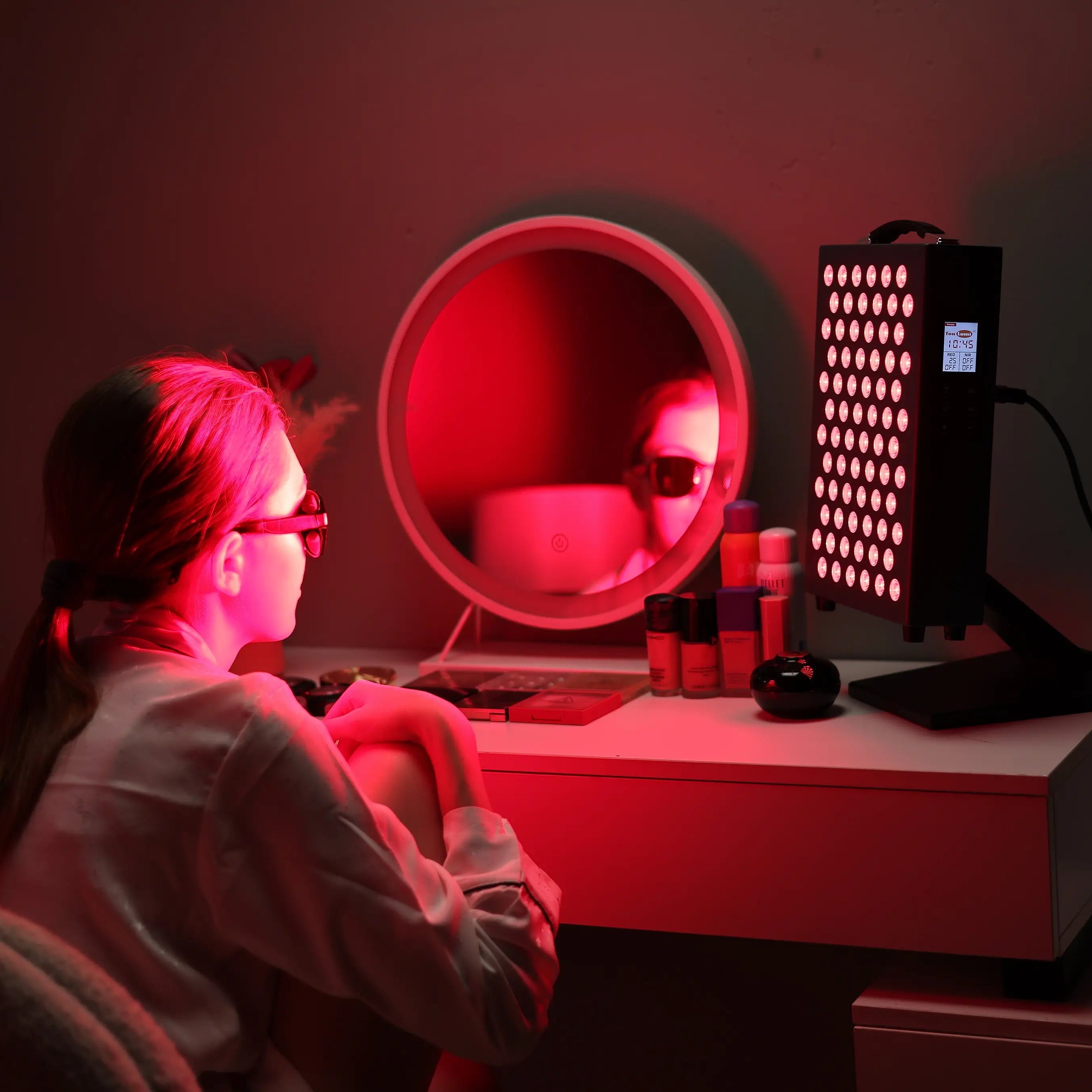 Woman using YouLumi red light therapy panel at a vanity, desktop unit with LEDs on