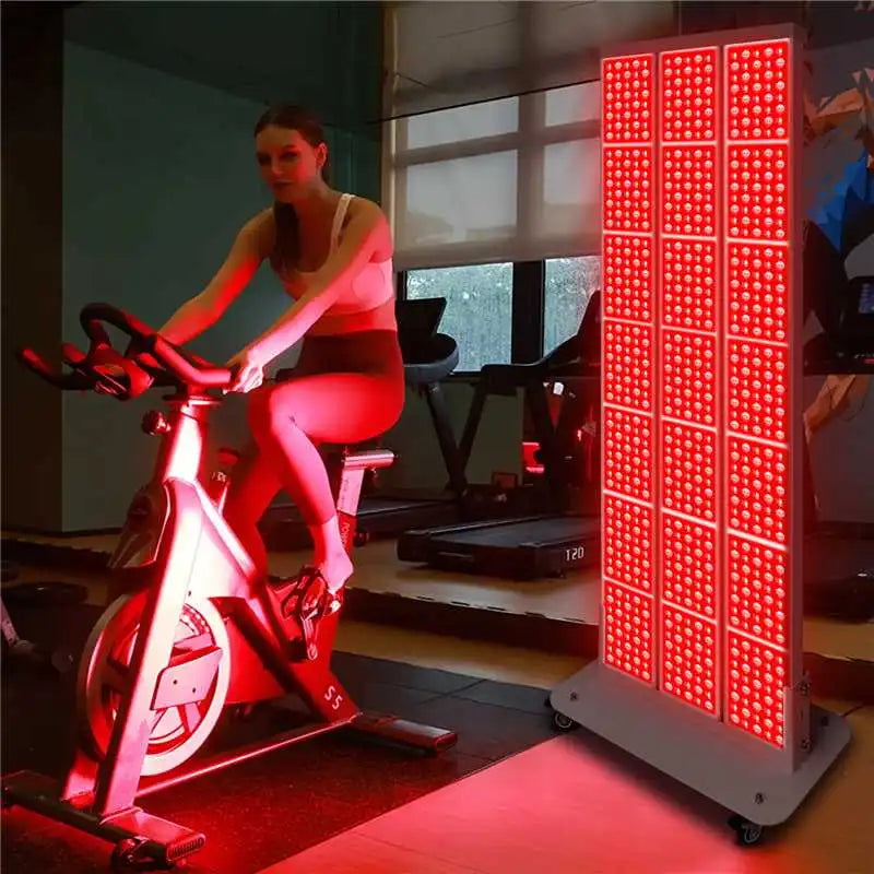 Woman cycling on a stationary bike in a gym while illuminated by a tall wheeled red light therapy panel