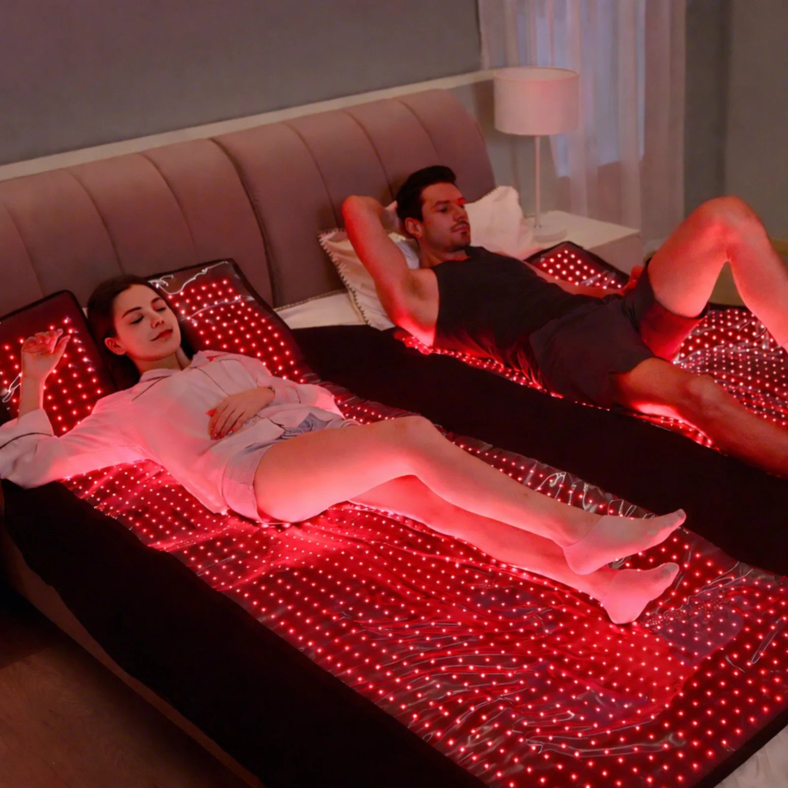 Couple relaxing on separate laser + LED sleep bag mats in a bedroom with full-body red light therapy glow.