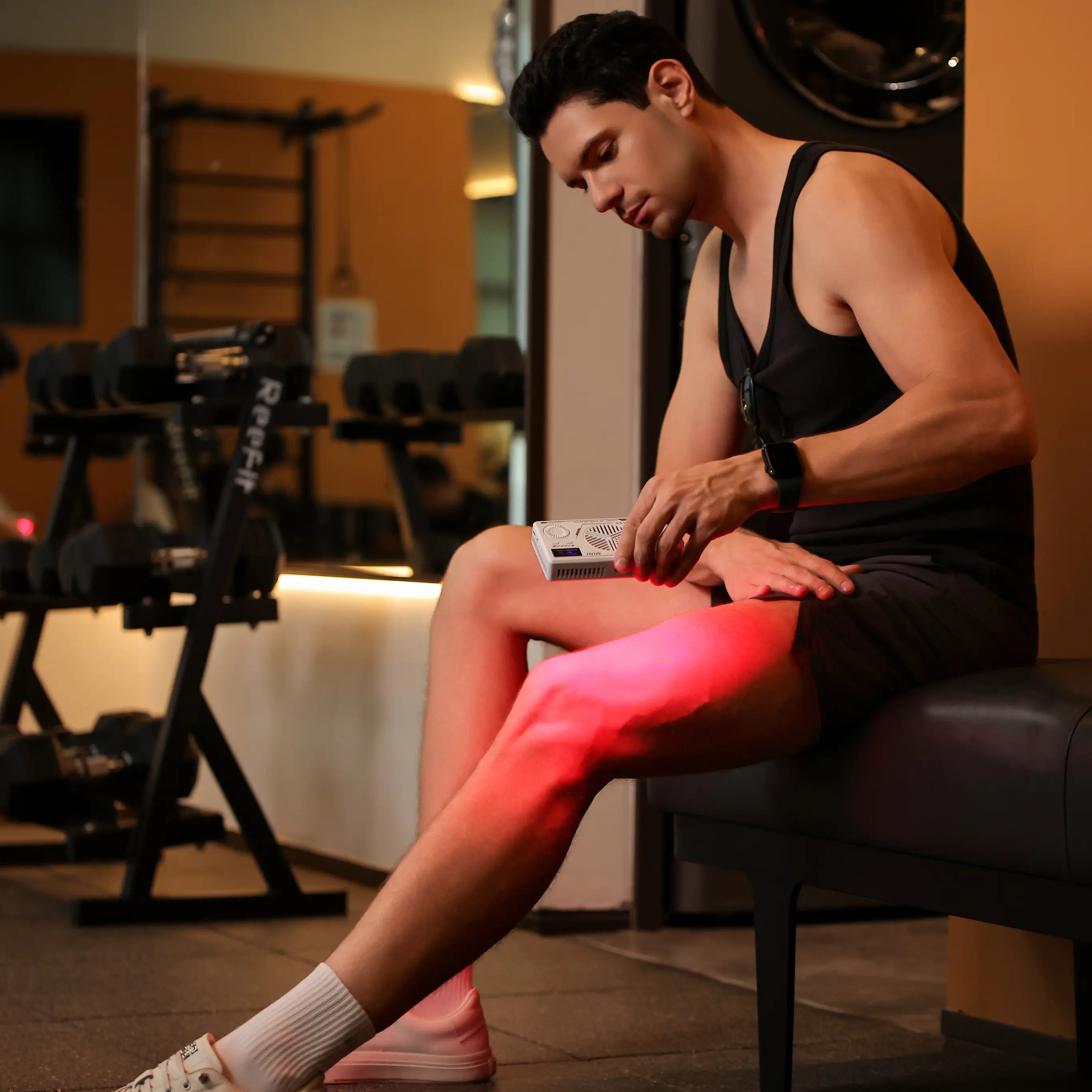 Athletic man sitting on a gym bench applying a red light therapy device to his knee and thigh for muscle recovery and pain relief after a workout.