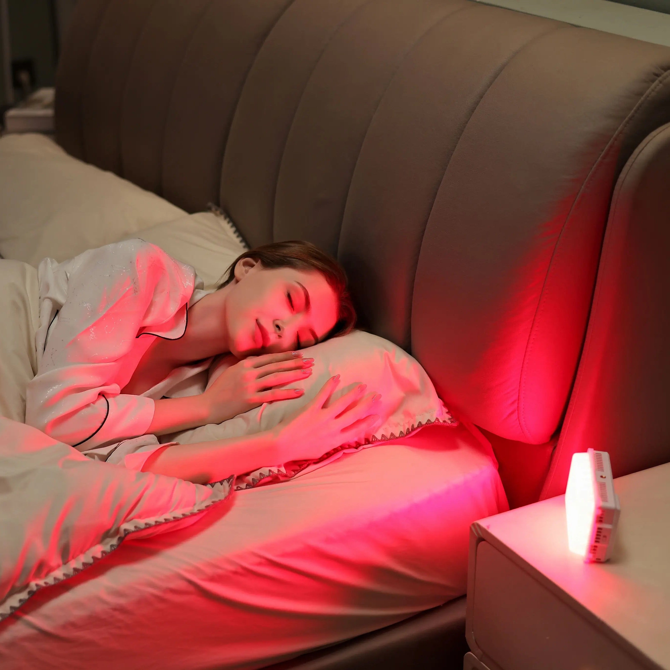 Woman sleeping peacefully in a comfortable bed with a red light therapy device placed on the nightstand emitting a warm glow to aid sleep.